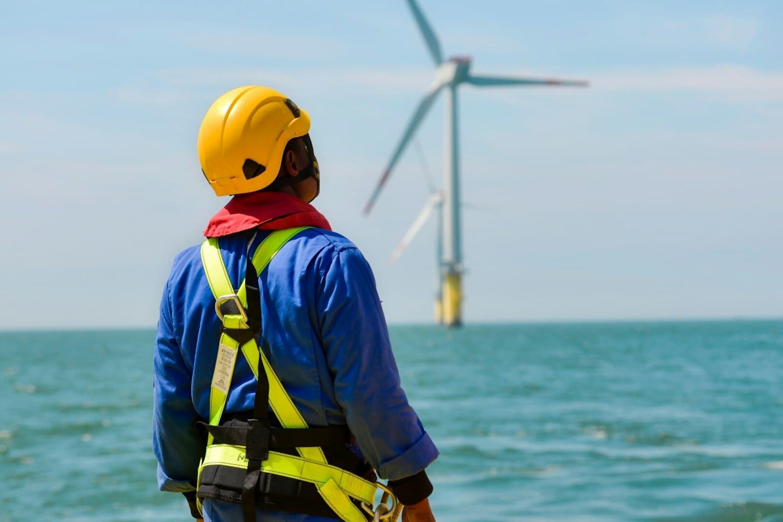 Beschäftigter mit Helm und Sicherheitsgurt blickt über das Wasser auf eine Offshore-Windenergieanlage